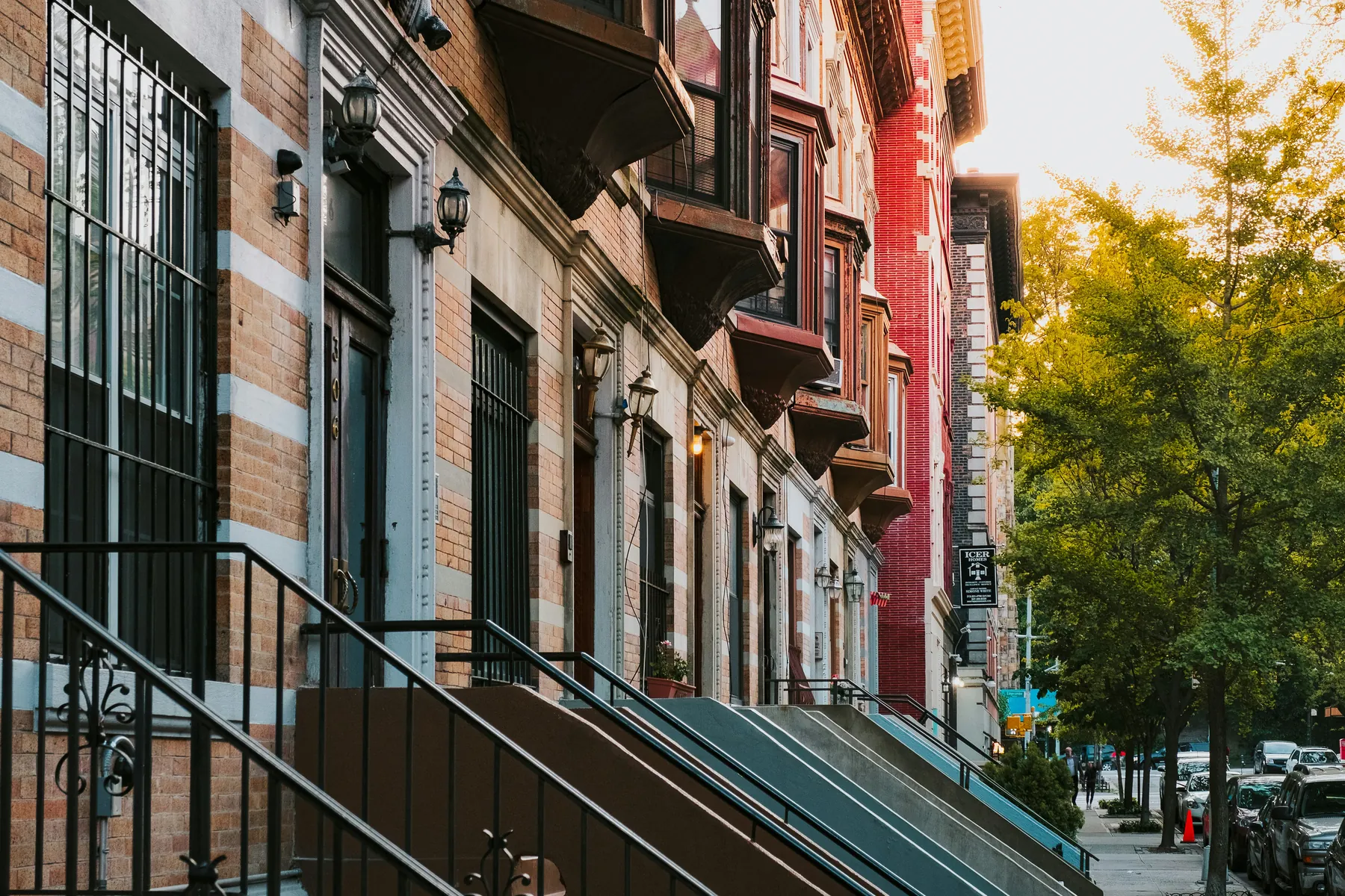 A row of brick and brownstone townhouses on a Harlem street, with black iron railings and a yellowing tree in autumn light