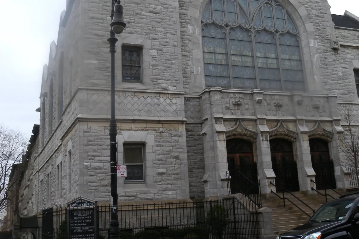 The grey stone Gothic front of a Harlem church, with a square tower, a large arched stained-glass window and three arched doorways