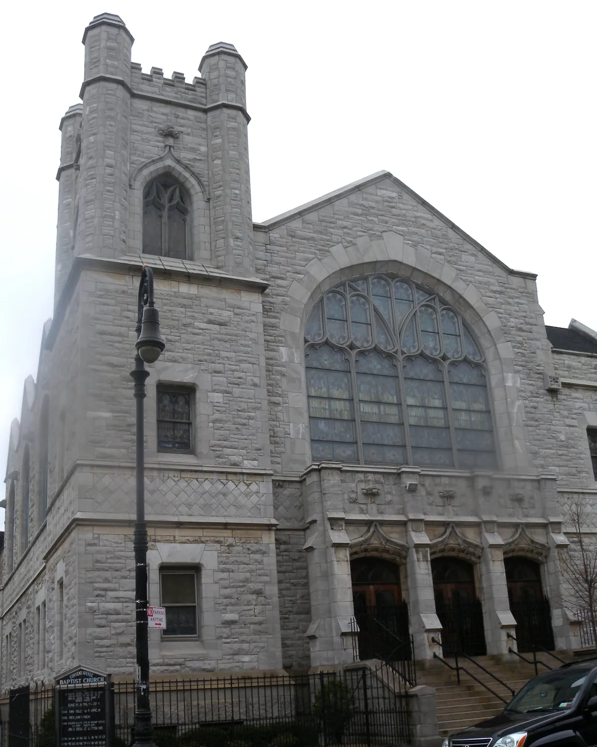 The grey stone Gothic front of a Harlem church, with a square tower, a large arched stained-glass window and three arched doorways