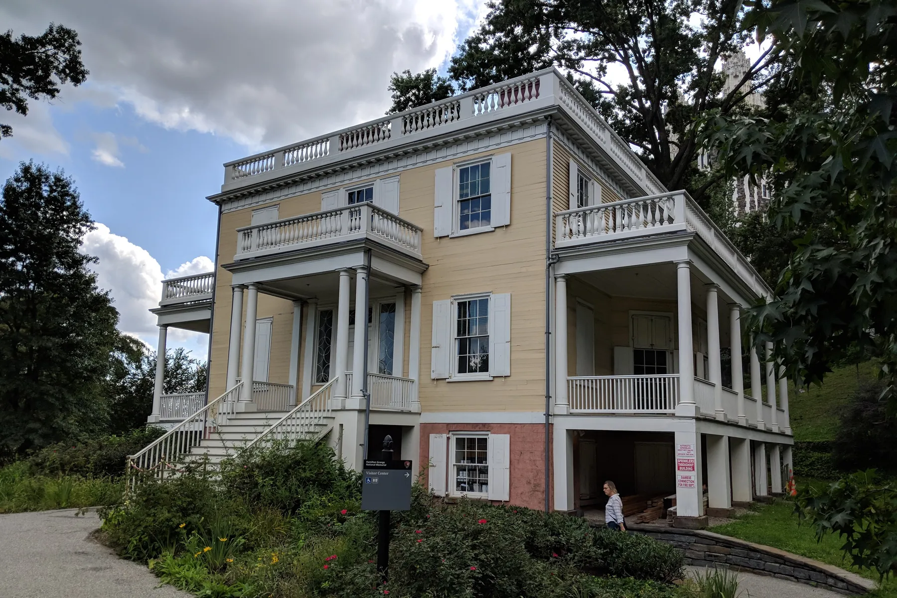 A pale yellow wooden house with white two-storey porches and balconies, set among trees on a curving path