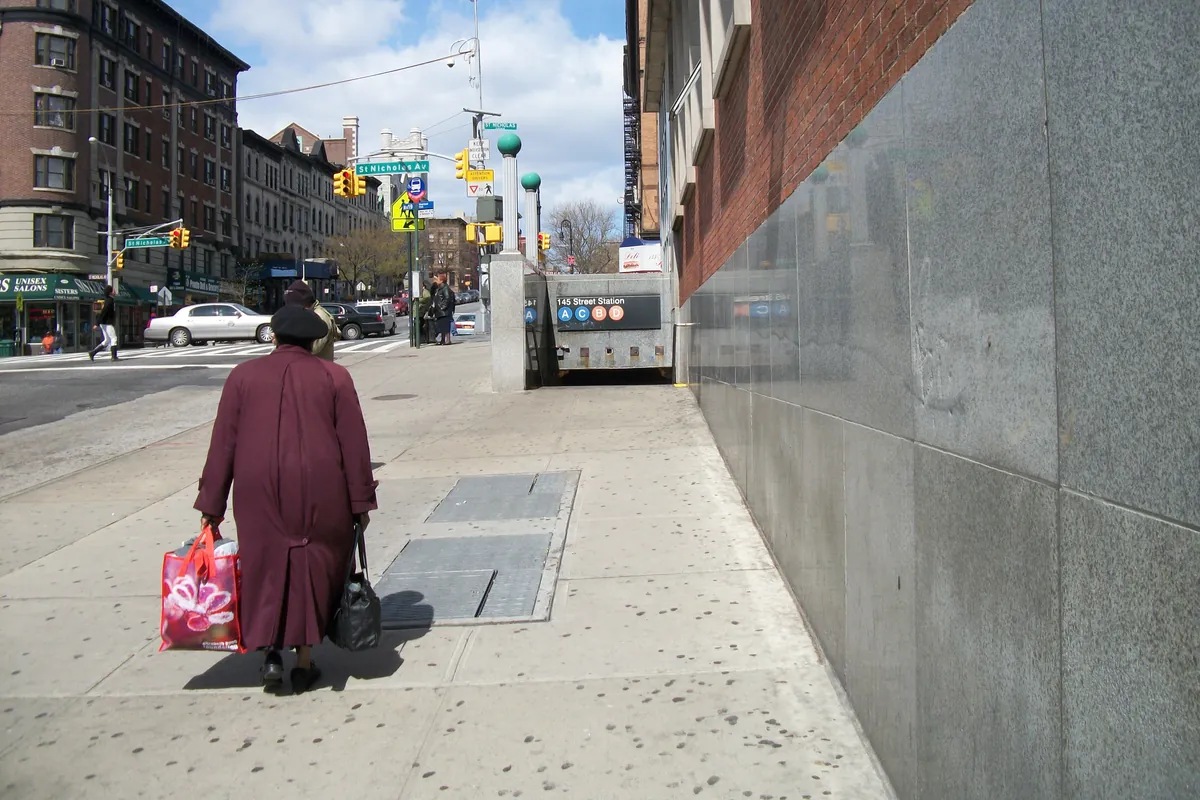 A subway entrance on a Harlem street corner, with a woman carrying shopping bags walking past tall brick apartment buildings